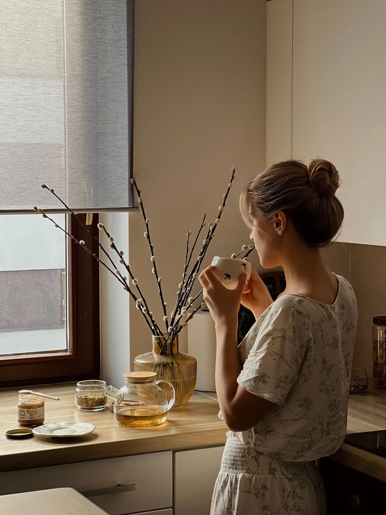 A woman standing at a sunlit kitchen window with herbal tea and pussy-willow branches